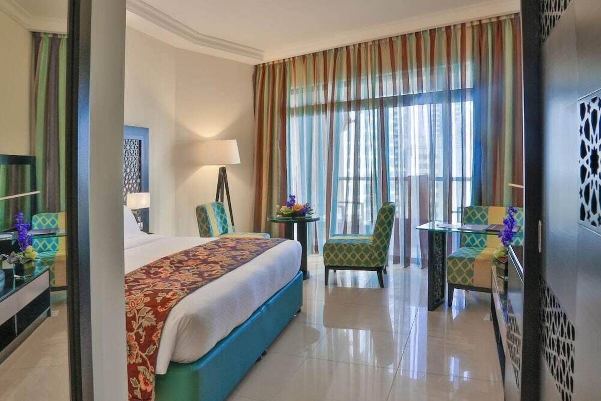 Habitación de hotel con cama, escritorio, silla y un amplio ventanal con cortinas.