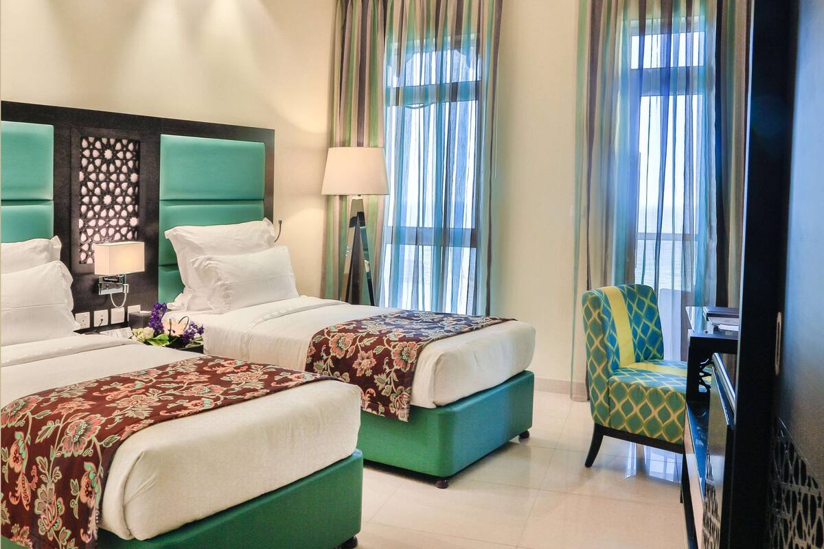 Una habitación de hotel con dos camas, un sillón verde, un escritorio y un ventanal con cortinas.