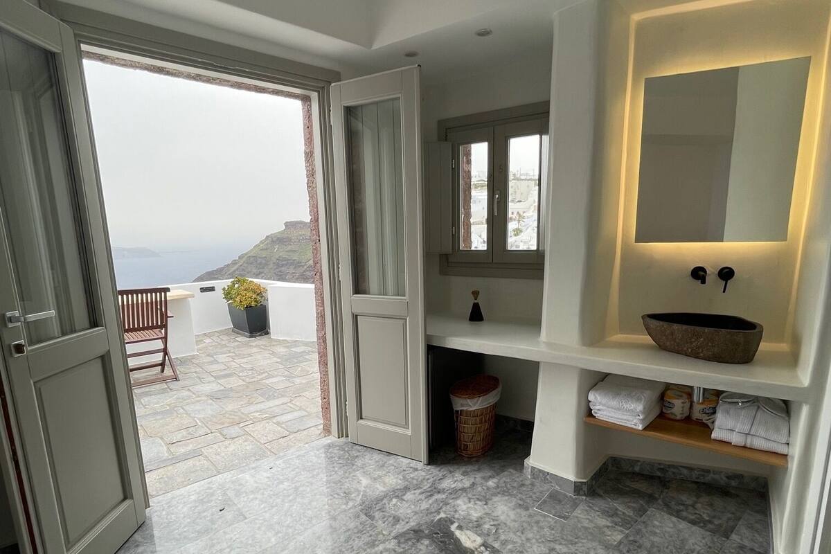 Un baño moderno con suelo de mármol, un espejo grande y vistas al mar a través de una puerta abierta.