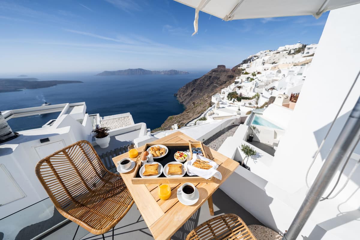 Un balcón con una mesa dispuesta para el desayuno, con vista a un paisaje costero de edificios blancos y el mar.