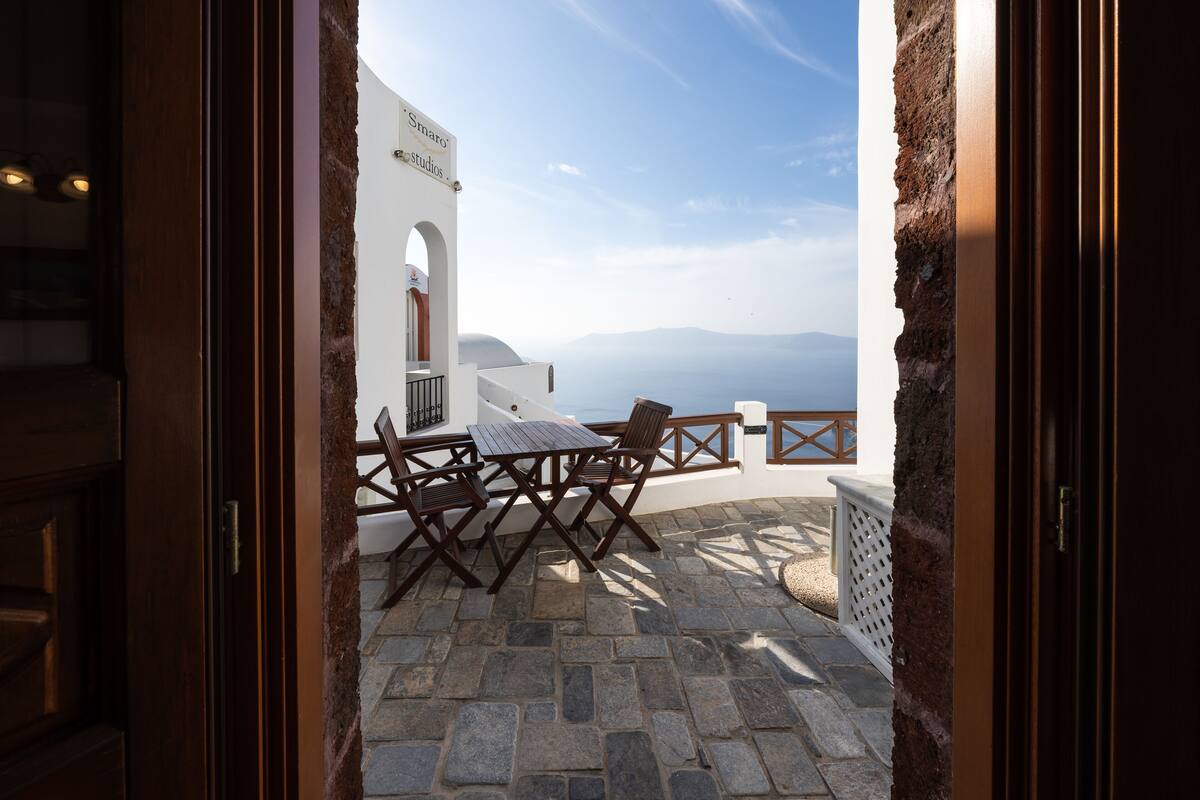Vista desde un vano de puerta a un patio exterior con suelo de piedra, una mesa y sillas de madera, y un edificio blanco con un cartel.