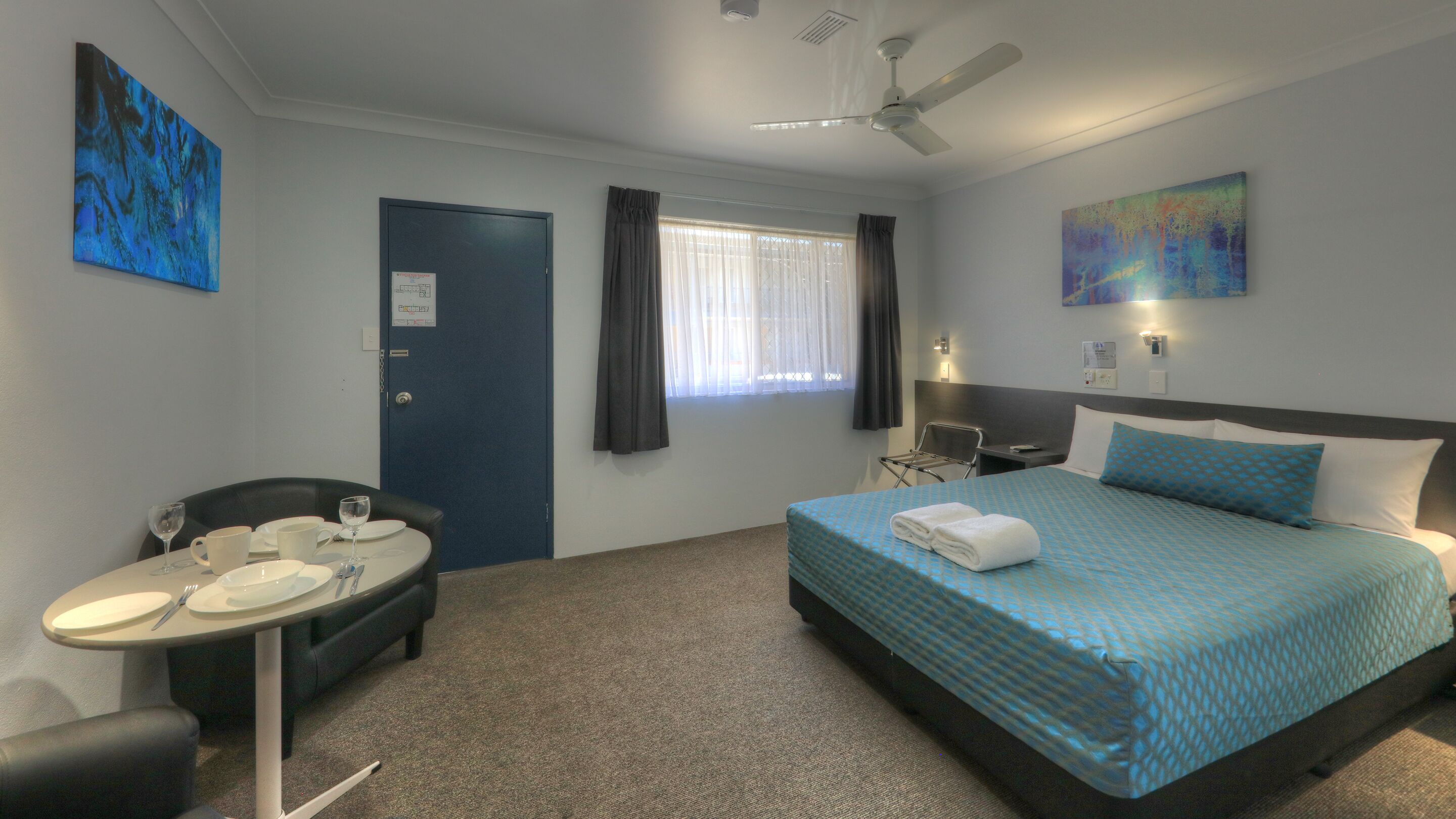 A hotel room with a bed, a desk with a chair, a dining table, and a window with curtains.