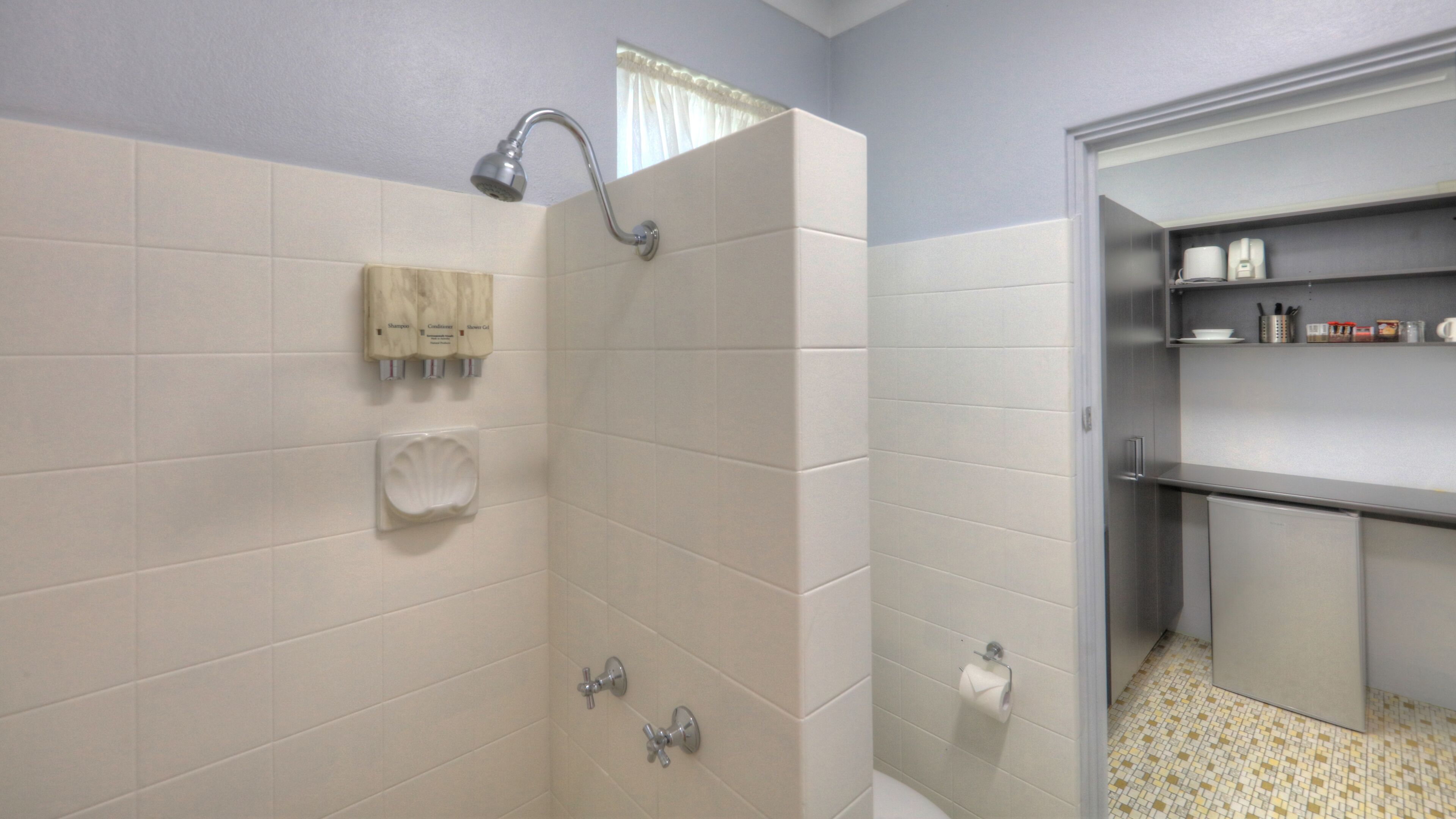 A bathroom with a shower, toilet, and sink. There are toiletries on the wall and a shelf with cups and containers.