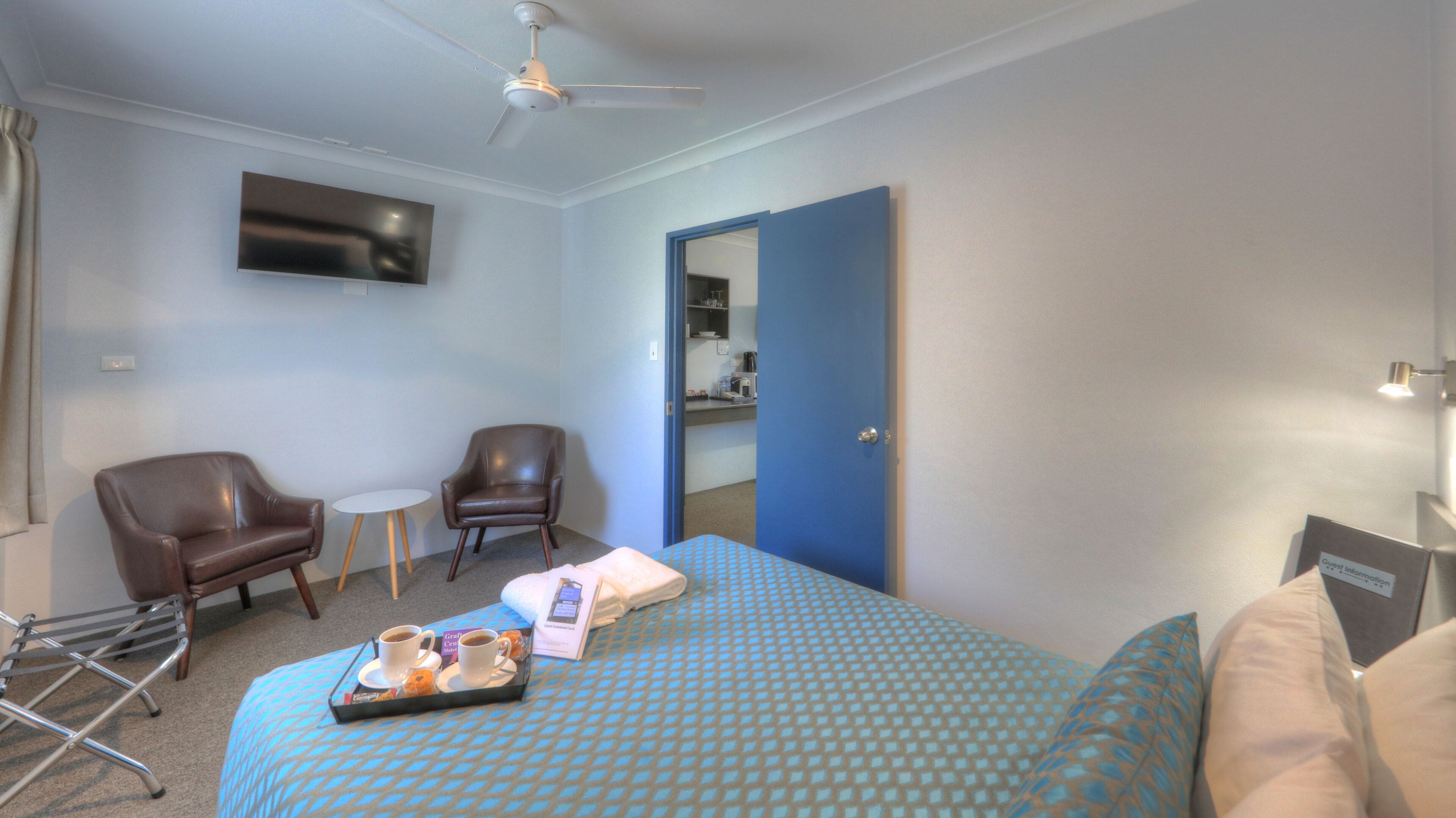 A hotel room with a bed, two chairs, a small table, a ceiling fan, a TV, and a bathroom visible through a door.