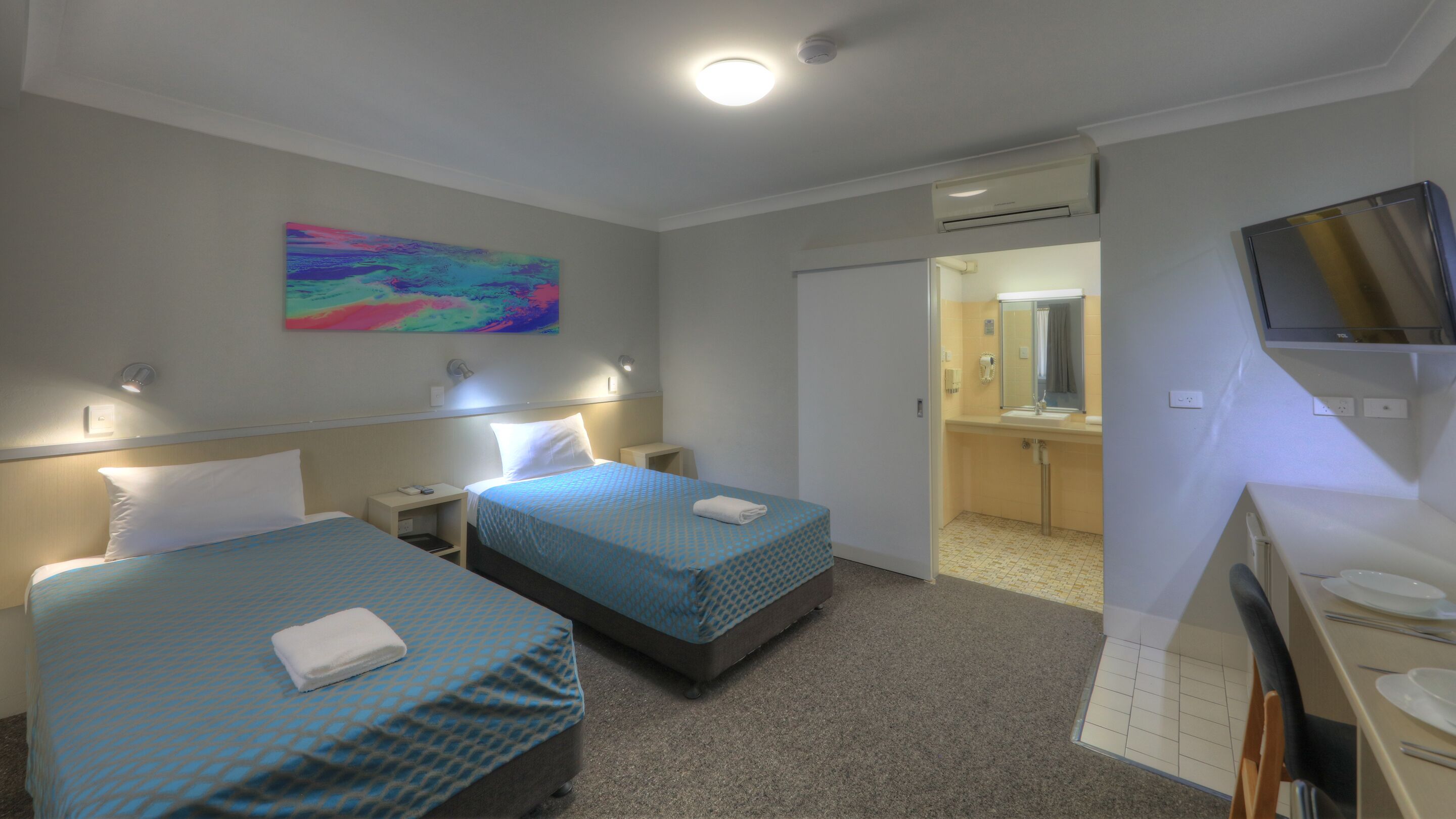 A hotel room with two beds, a TV, a bathroom, and a painting on the wall.