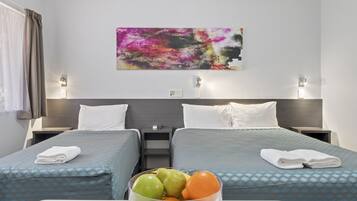 Un cuarto de hotel con dos camas, una mesita de noche con lámpara, un cuadro abstracto en la pared y un frutero sobre la mesita.