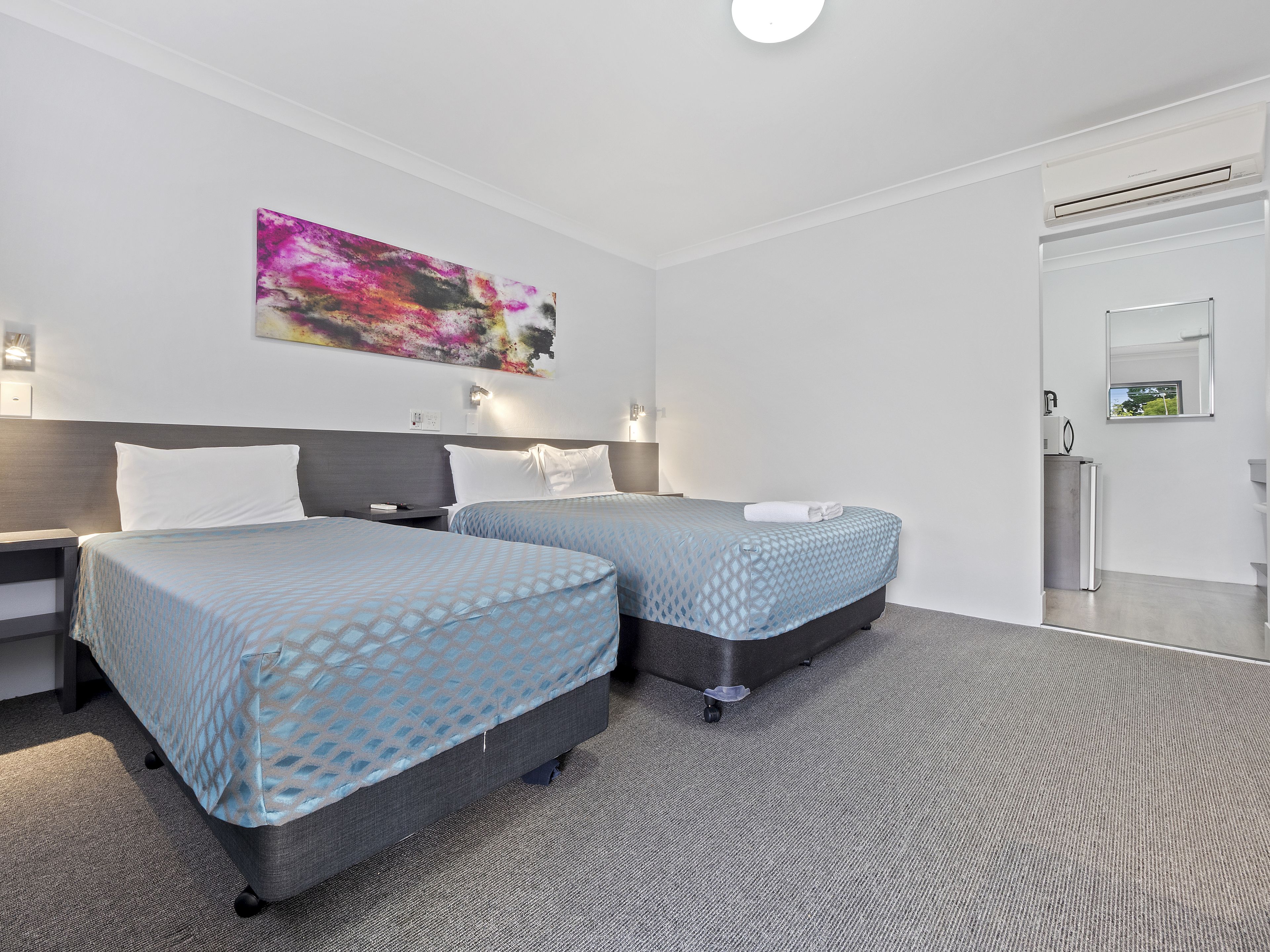 A modern hotel room with two beds, a large abstract painting, a small desk, and a compact kitchenette.