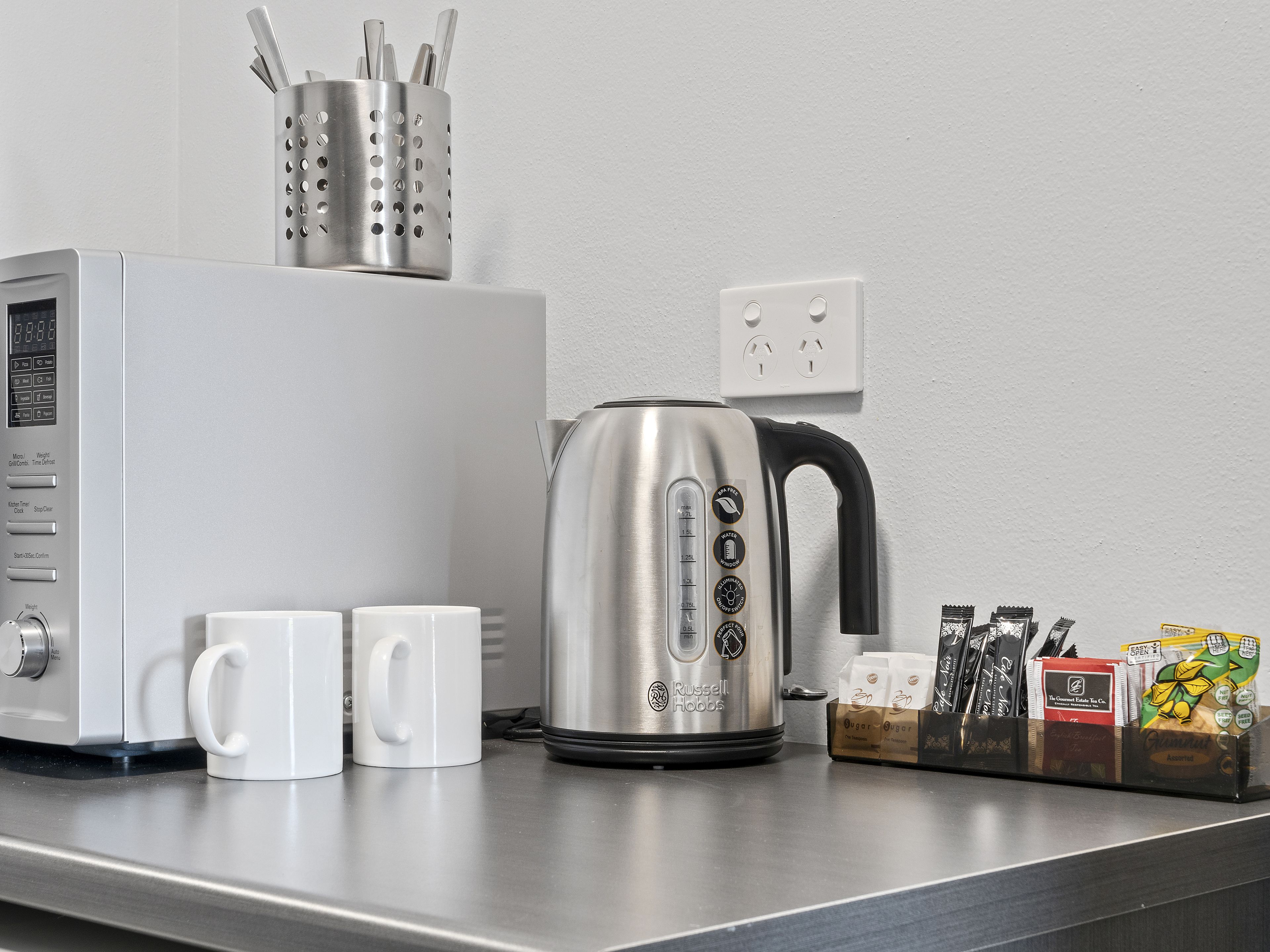 Una zona de cocina con cafetera, dos tazas blancas, un hervidor y un soporte para cápsulas de café con diferentes marcas.