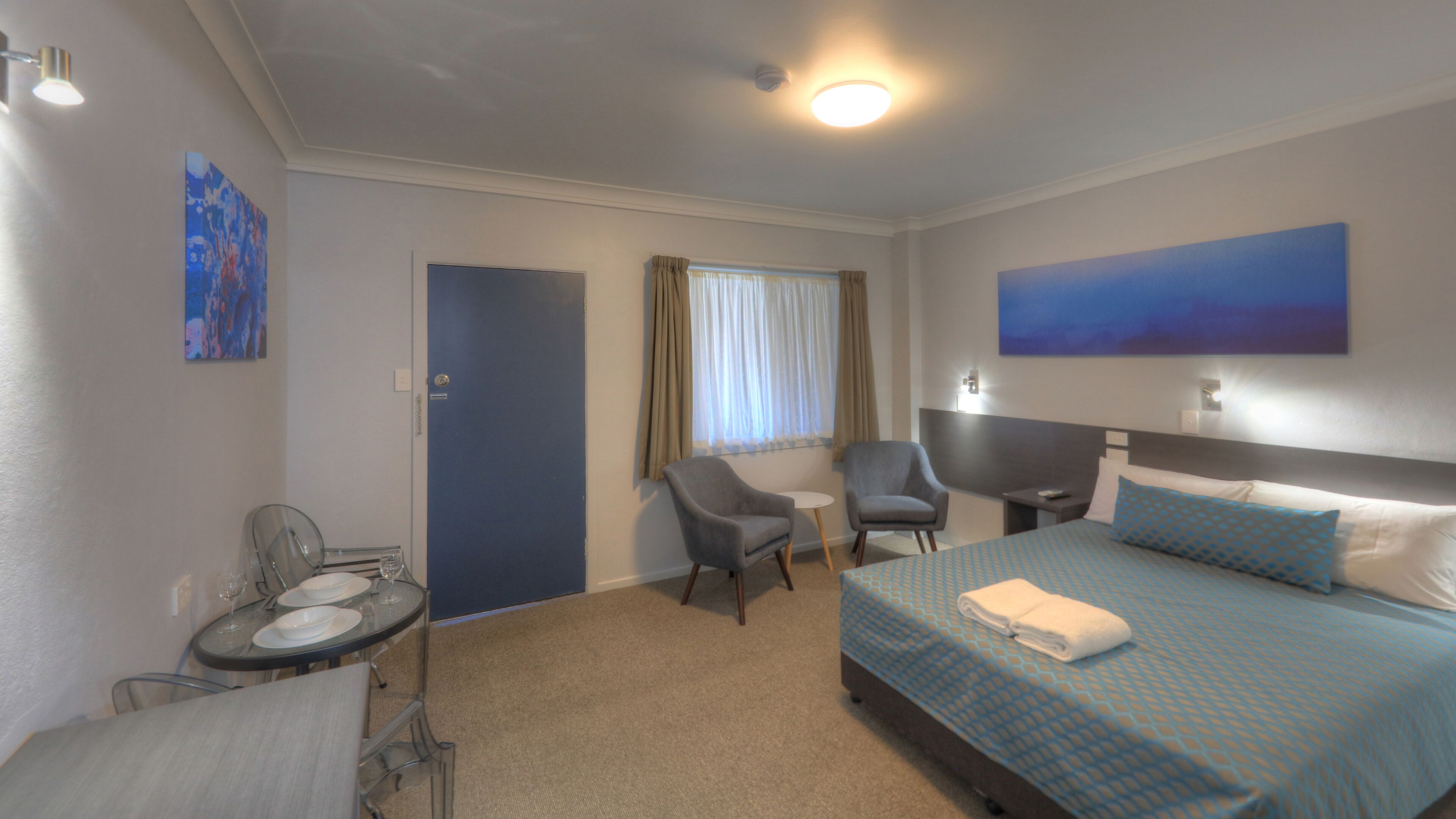 Habitación de hotel con cama, dos sillones, una mesita con un vaso y un cuadro en la pared.