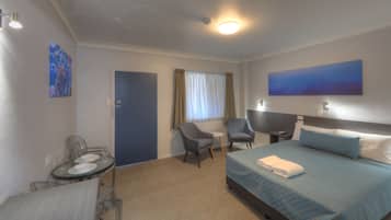 Habitación de hotel con cama, dos sillones, una mesita con un vaso y un cuadro en la pared.