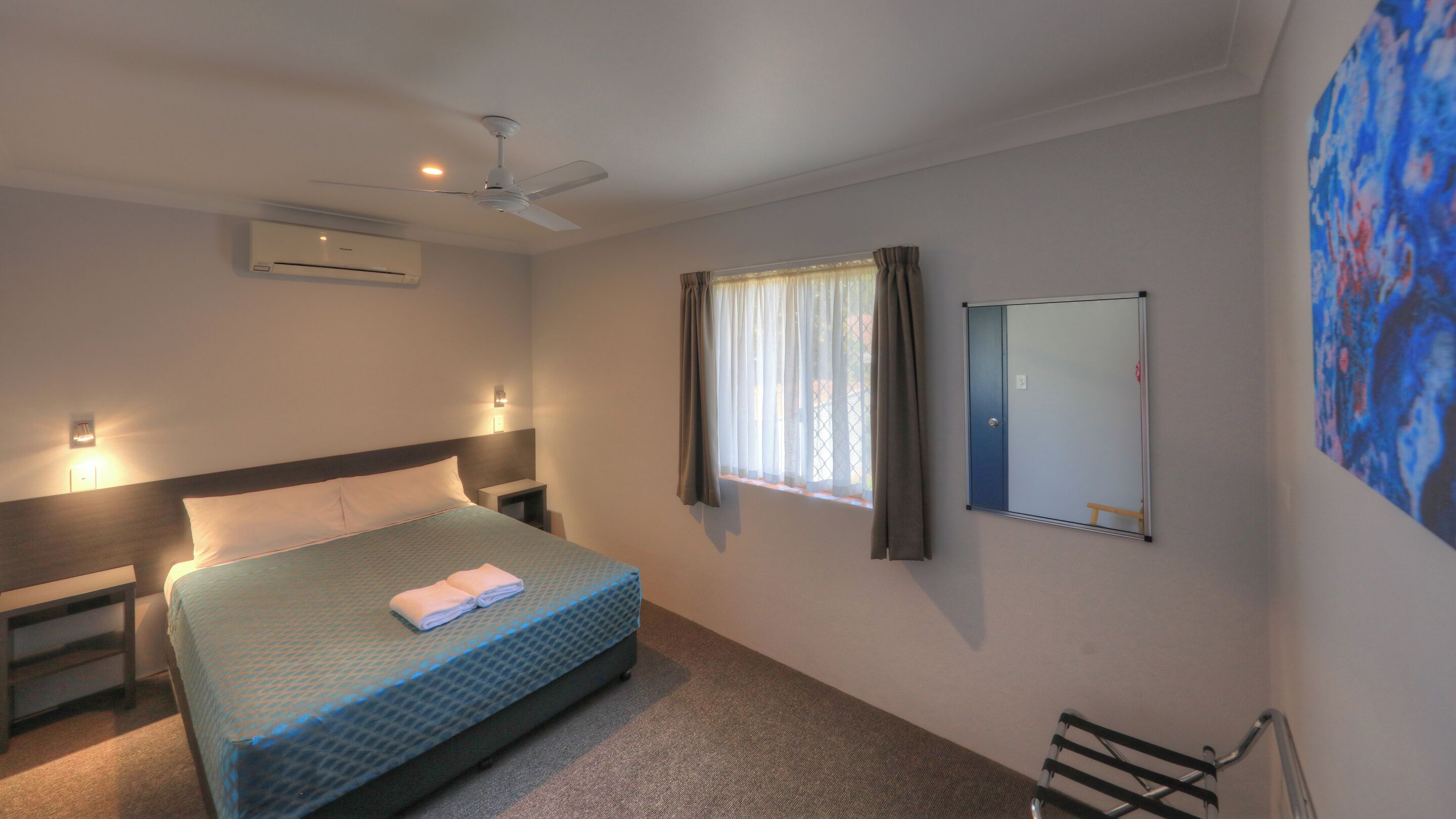 Habitación de hotel con cama, ventilador de techo, unidad de aire acondicionado, mesita de noche y espejo montado en la pared. Tiene además una ventana con cortinas.