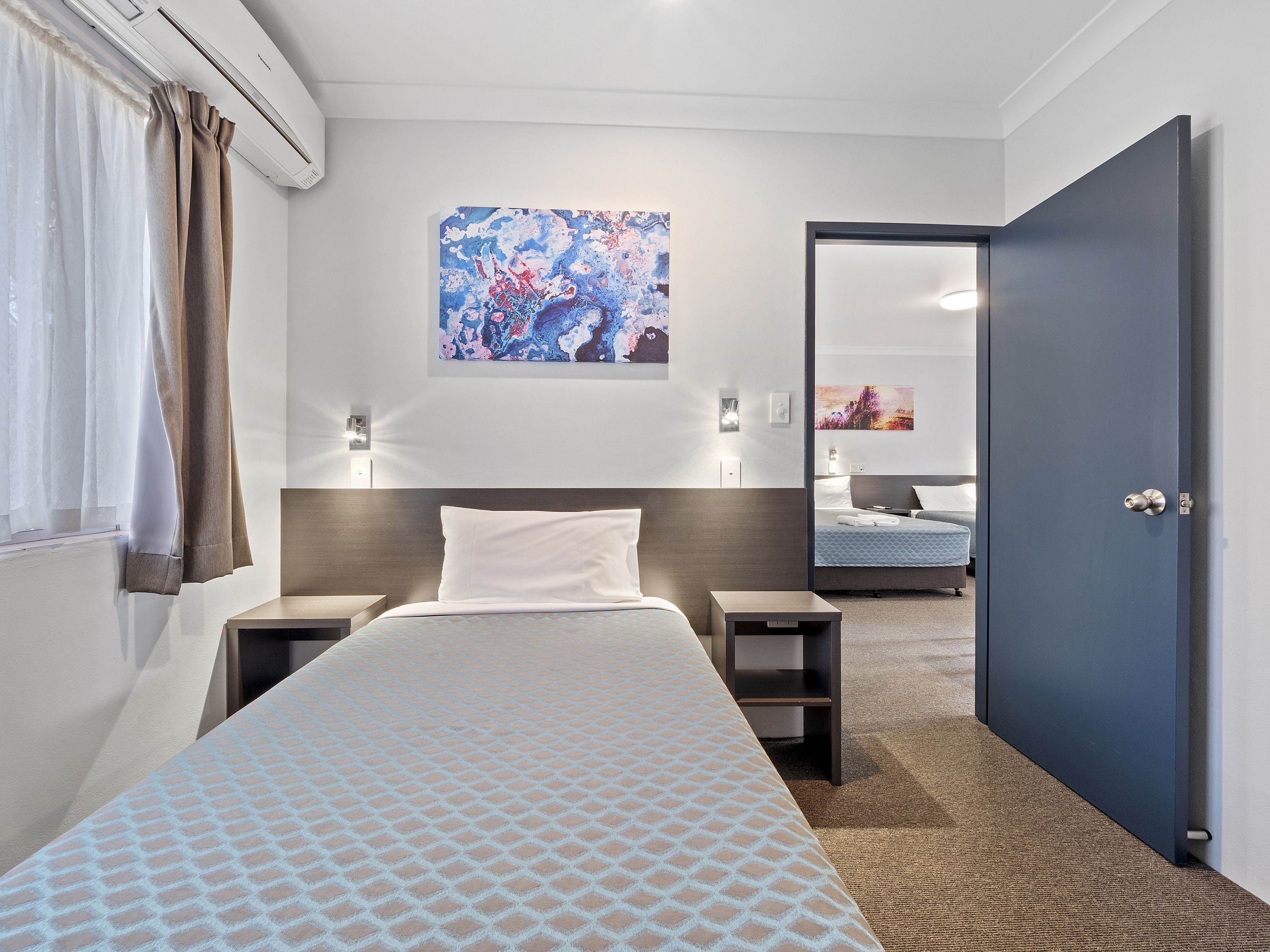 A hotel room with a bed, bedside tables, a nightstand, a wall-mounted artwork, a door, and a carpeted floor.