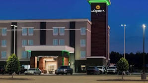 Hotel La Quinta con diseño moderno, que presenta una torre destacada con el logotipo del hotel, una entrada bien iluminada y un estacionamiento con varios automóviles.