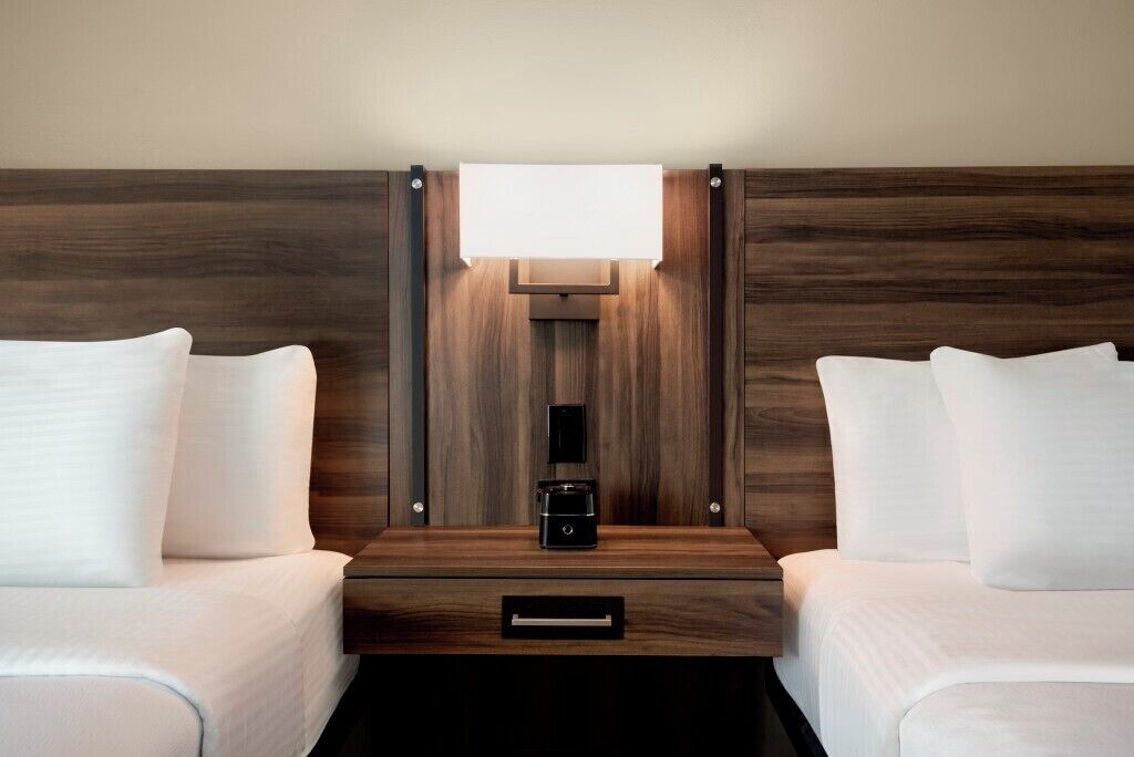 A hotel room with two beds, a bedside table with a coffee maker, and a wall-mounted light fixture.