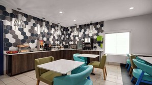Una cafetería moderna con paredes de diseño hexagonal, una barra con máquinas de café y sillas color turquesa alrededor de mesas blancas.