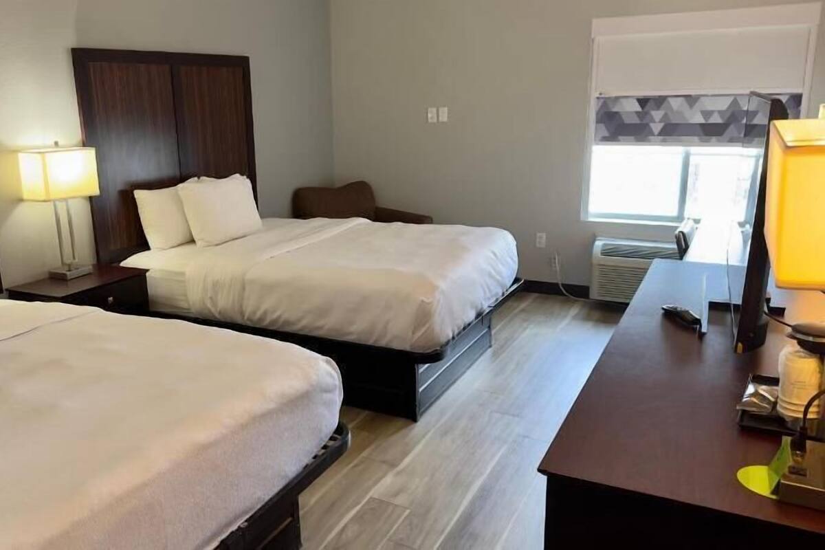 A hotel room with two beds, a desk, a chair, a lamp, and an air conditioning unit.