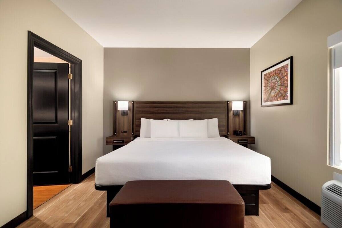 A hotel room with a large bed, bedside lamps, a wooden headboard, a small table, and a door.