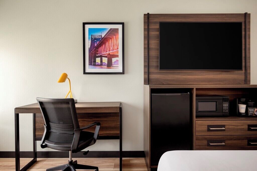 Une chambre d’hôtel équipée d’un bureau, d’une chaise, d’un lit, d’une télévision, d’un four à micro-ondes et d’un tableau encadré au mur.