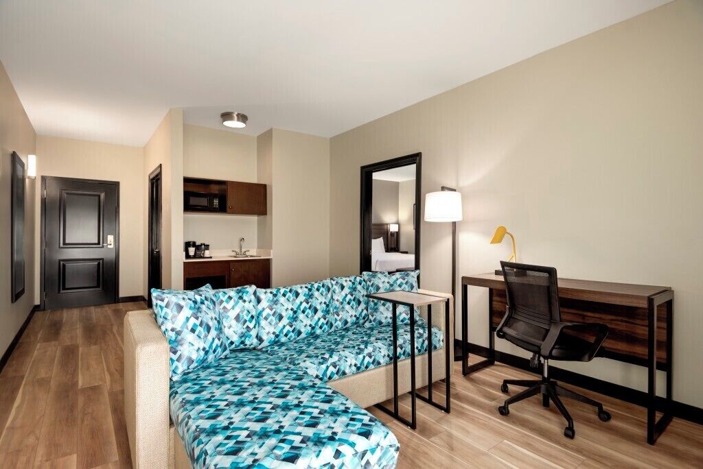A modern hotel room with a sectional sofa, a desk with a chair, a mirror, and a small kitchenette.