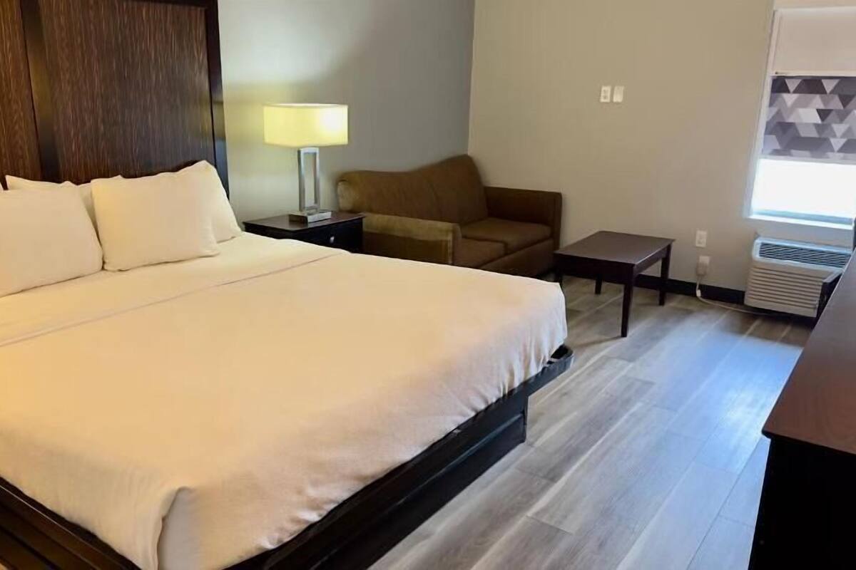 A hotel room with a large bed, a sofa, a small table, a lamp, and an air conditioning unit.