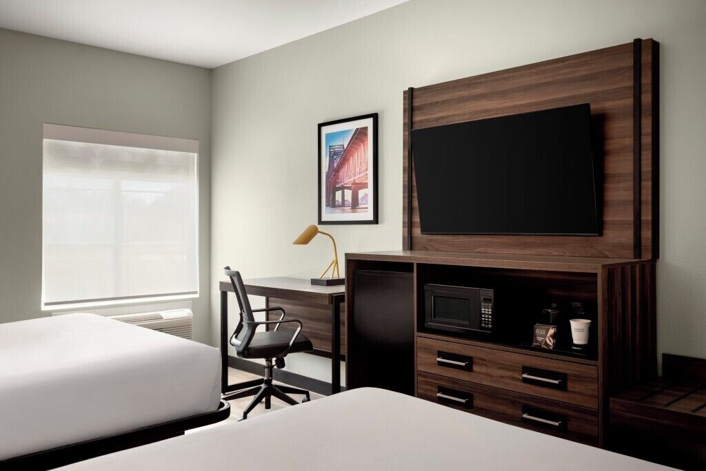 Quarto de hotel com uma cama, uma escrivaninha, uma cadeira, uma televisão, um micro-ondas e um quadro na parede.