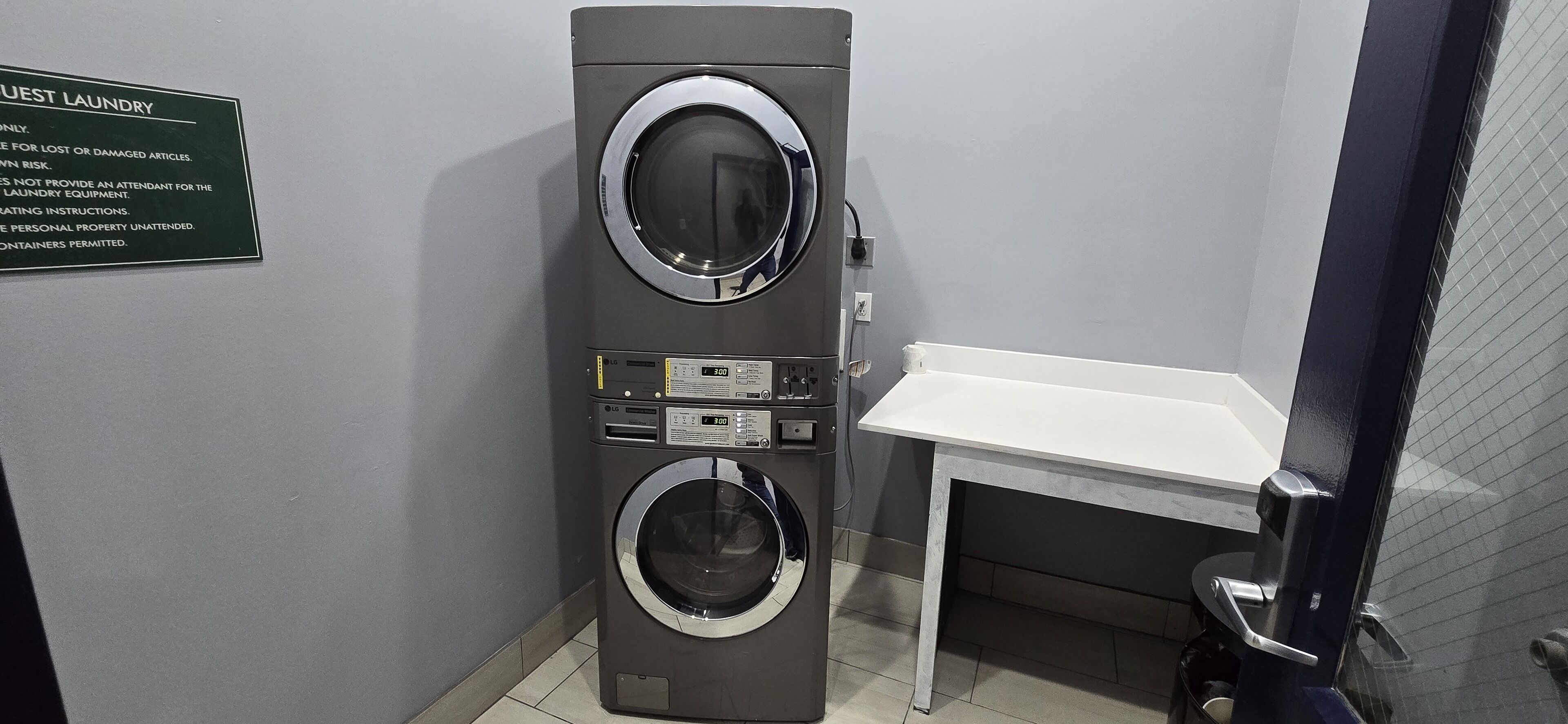 Una lavadora compacta de dos puertas con pantalla digital, una pequeña mesa y un letrero que indica uso para huéspedes.