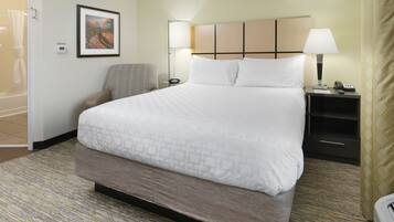 Una habitación de hotel con una cama grande, una mesita de noche con lámpara, una silla, un cuadro enmarcado en la pared y un termostato.