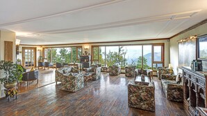 A spacious room with wooden flooring, floral-patterned sofas, and large windows offering a view of the outdoors. - Club Mahindra Mashobra (Shimla)