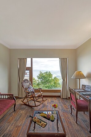Ein Zimmer mit Holzboden, einem Schaukelstuhl, einem Esstisch mit Lampe und Blick ins Grüne durch das Fenster.