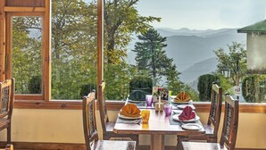 A dining area with a wooden table and chairs, a clock on the wall, and a view of mountains through large windows with bamboo blinds. - Club Mahindra Mashobra (Shimla)