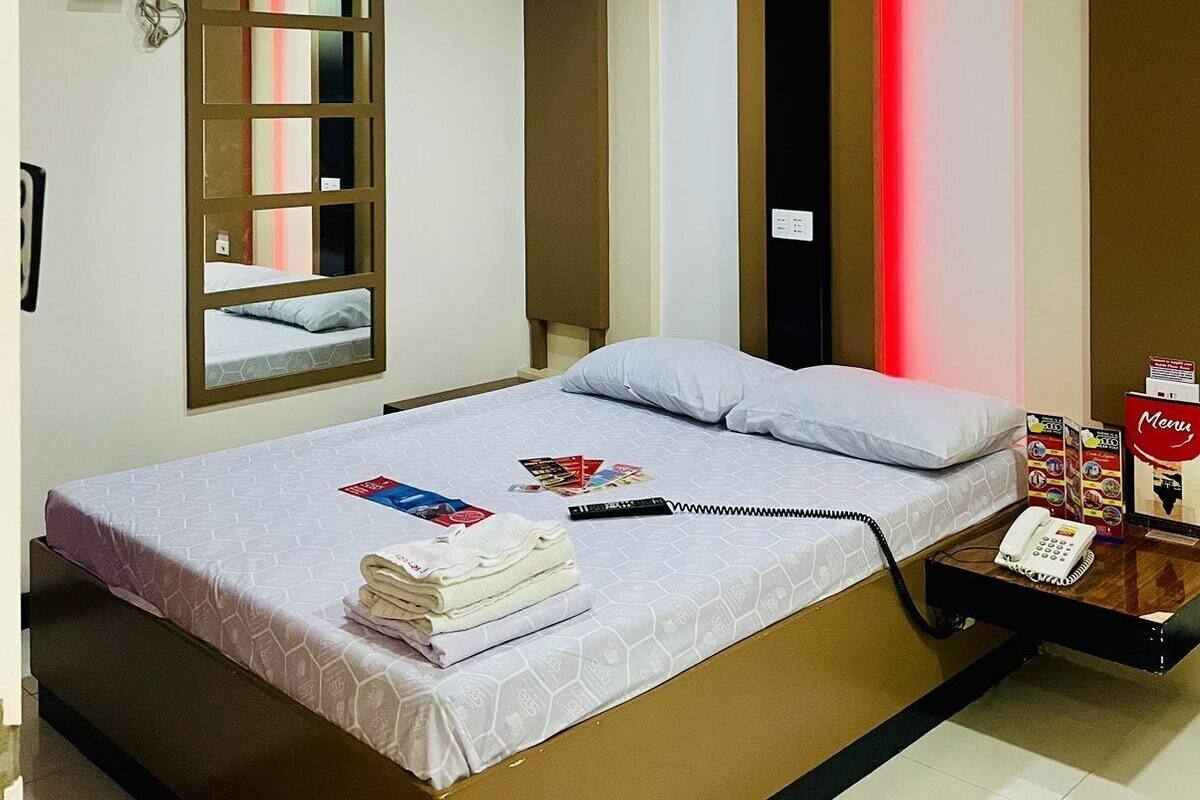 Une chambre d’hôtel avec un lit, une table de chevet avec un téléphone, un miroir et une fenêtre avec des stores.