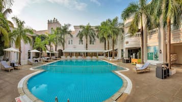 A pool area with lounge chairs and umbrellas, surrounded by palm trees and a building with arched windows.