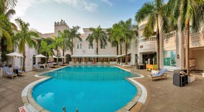 A pool area with lounge chairs and umbrellas, surrounded by palm trees and a building with arched windows.
