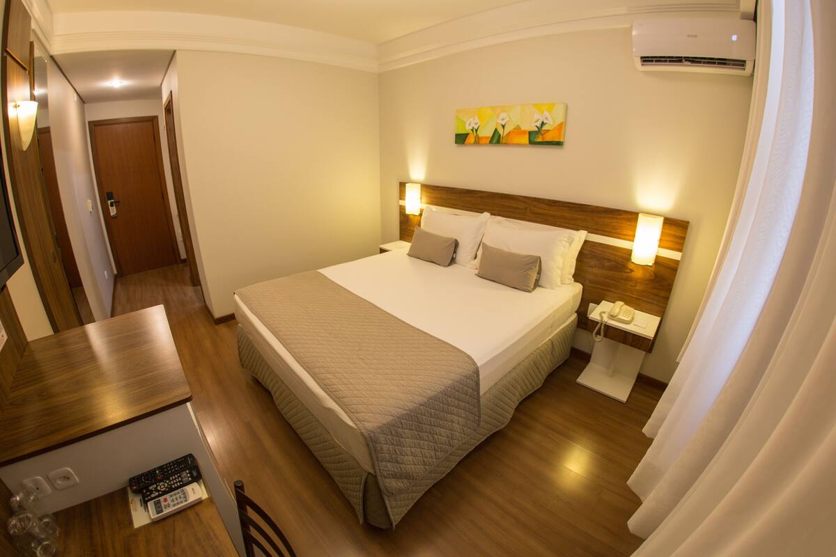 A hotel room with a large bed, bedside tables, a desk, and an air conditioning unit.