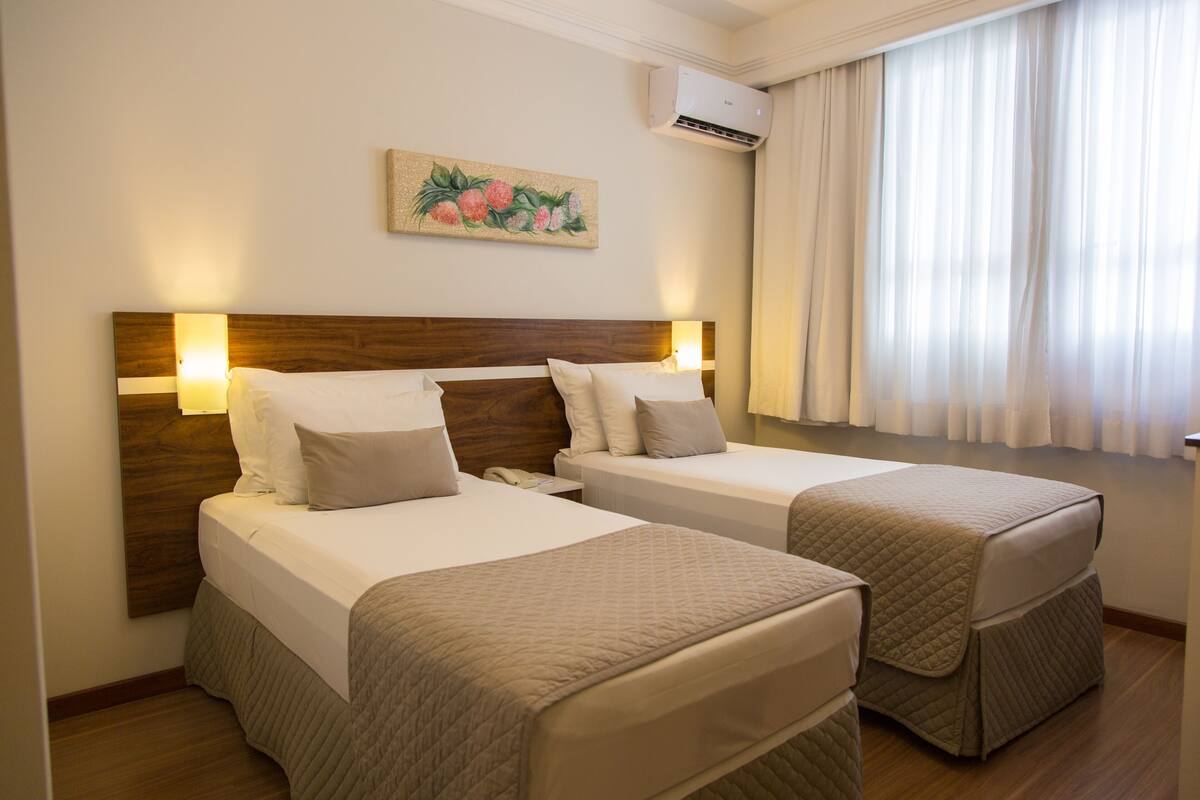 A hotel room with two beds, a wooden headboard, a painting, an air conditioning unit, and a window with curtains.