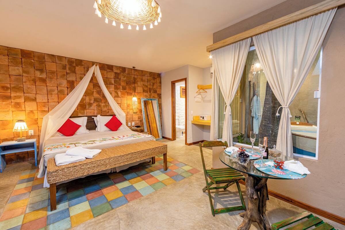 A bedroom with a four-poster bed, a small table with a vase and wine glasses, and a view of the sea through large windows.