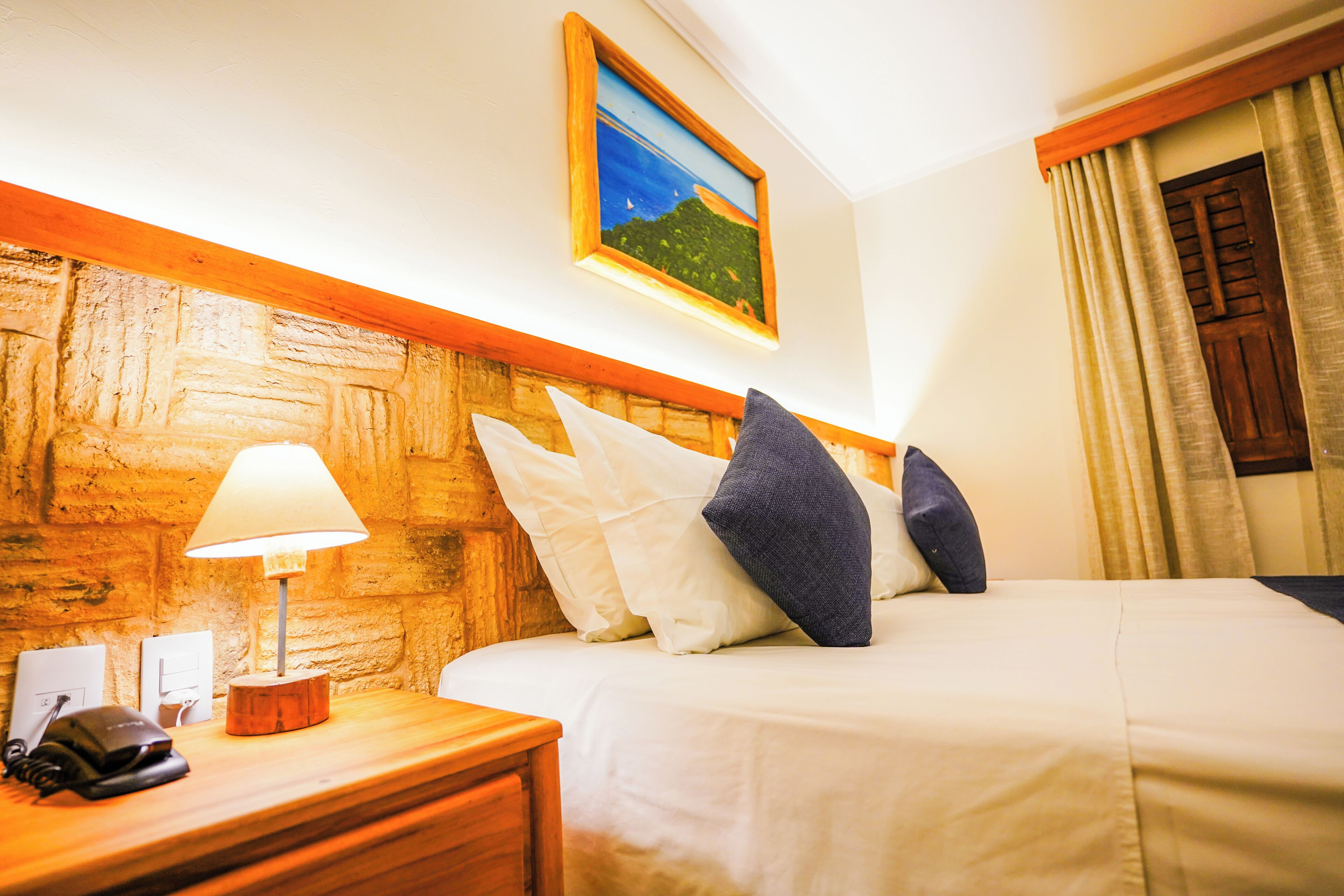 A hotel room with a wooden headboard, a bed with white and dark blue pillows, a bedside lamp, a telephone, and a painting on the wall.