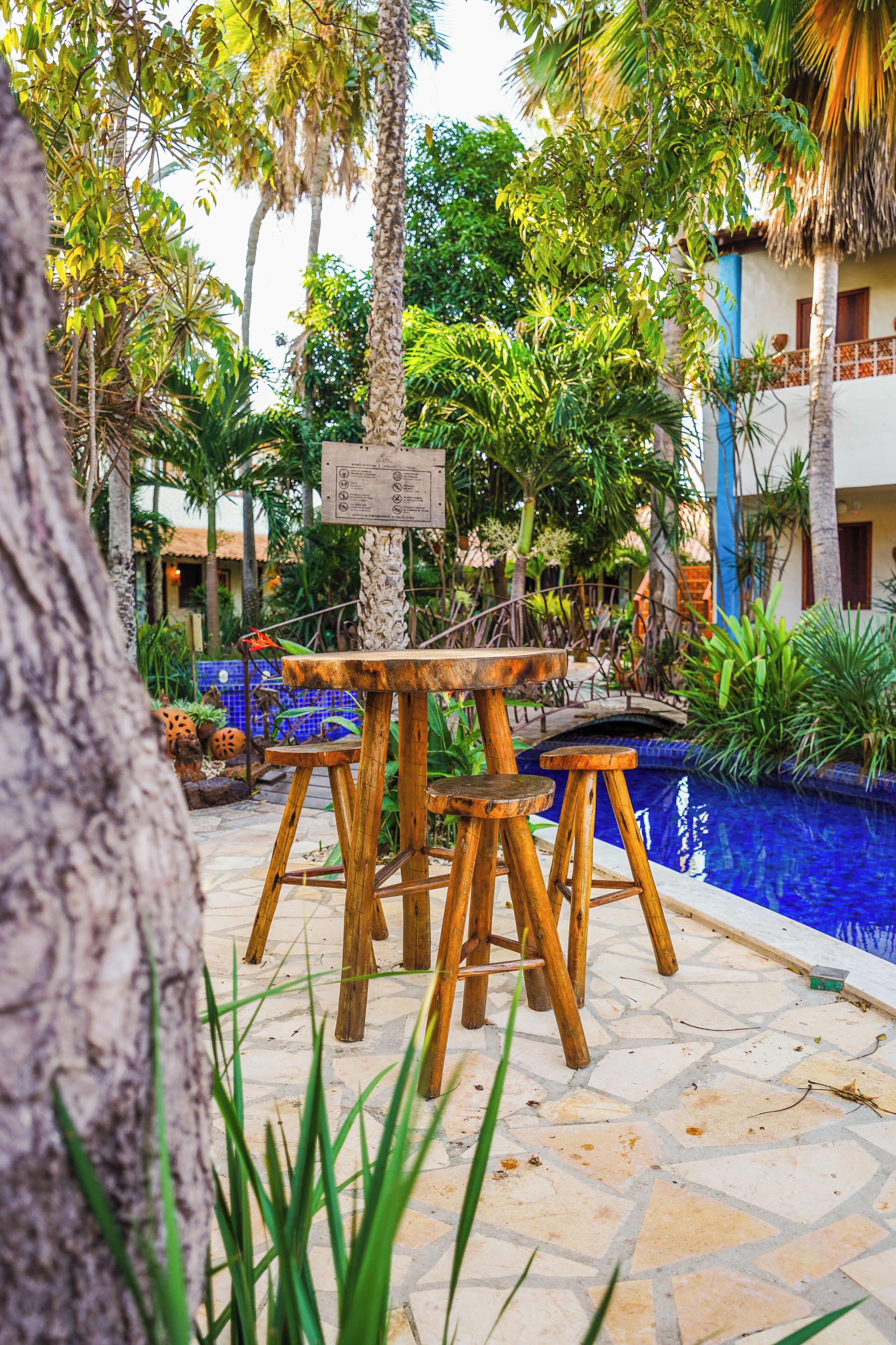 A poolside area with a wooden table and stools, surrounded by lush greenery and a clear blue pool.