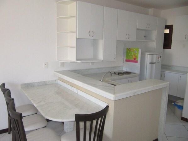Uma cozinha compacta com armários brancos, bancada de mármore e um pequeno espaço de refeições com uma cadeira.