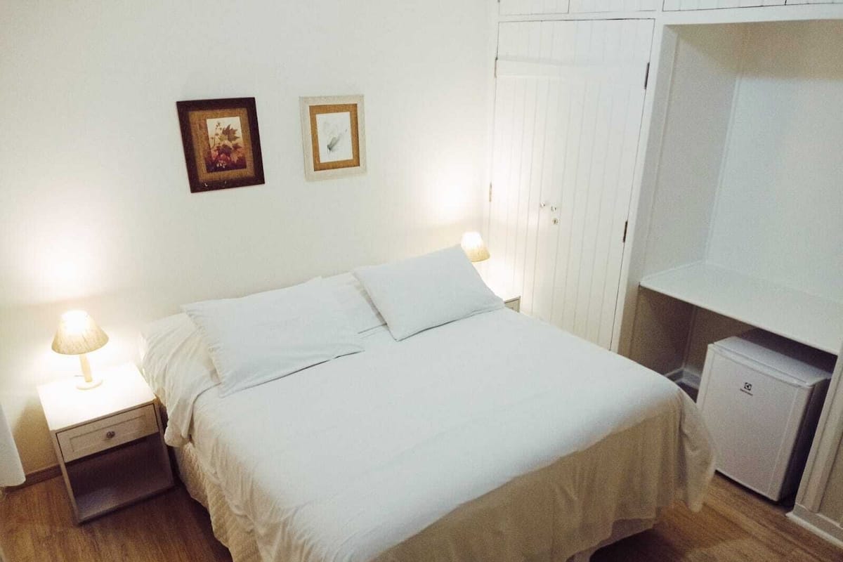 Una cama bien hecha con sábanas blancas, dos lámparas de noche, una mesita auxiliar pequeña y un armario con espejo.
