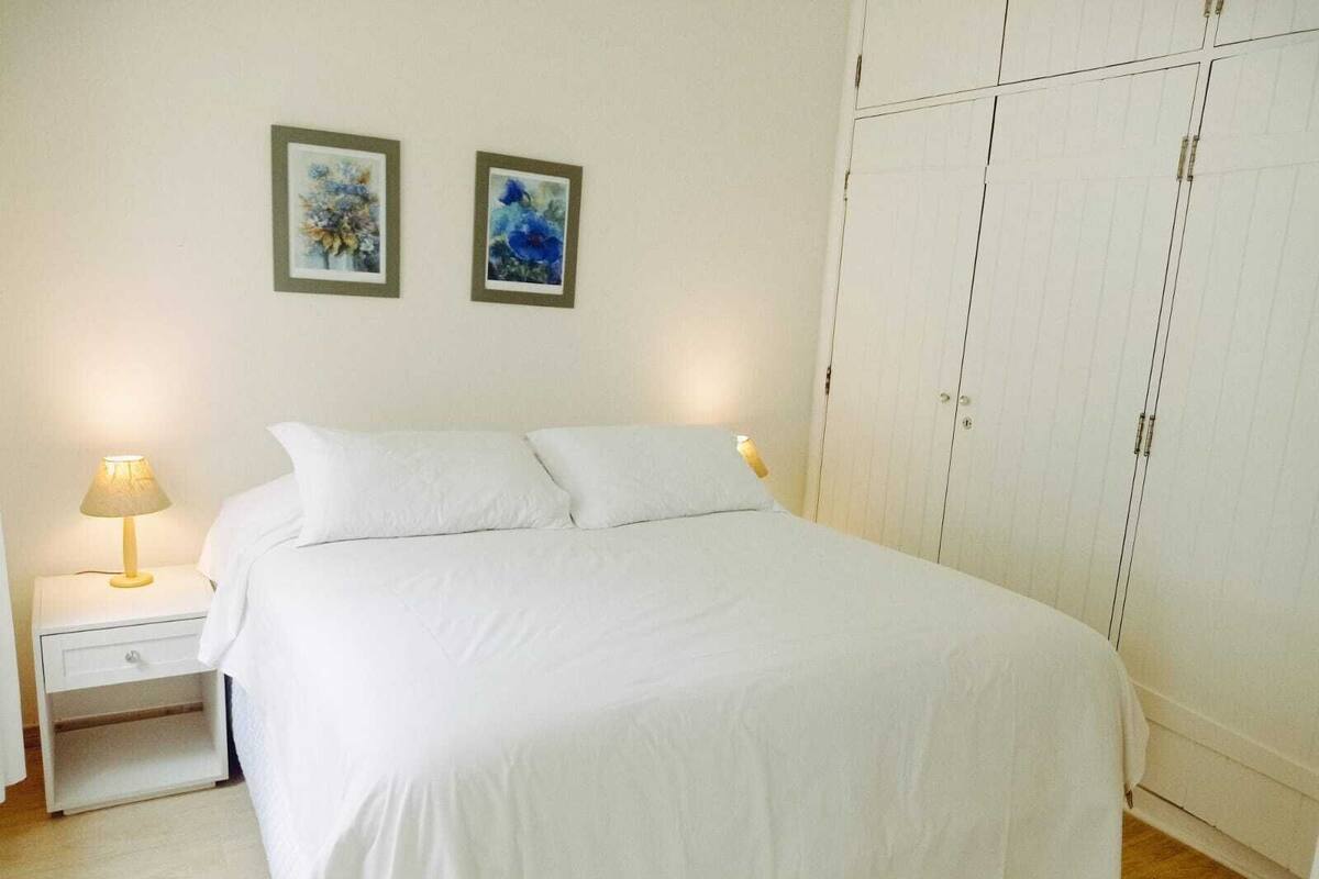 Una cama bien hecha en una habitación de hotel, con una colcha blanca, dos almohadas, una lámpara de noche y un armario con espejo.