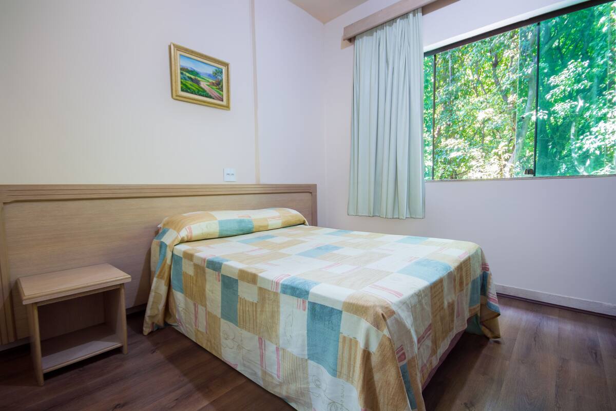 Ein Schlafzimmer mit einem Holzbett, einem kleinen Nachttisch, einem Fenster mit Vorhängen und einem gerahmten Bild an der Wand.