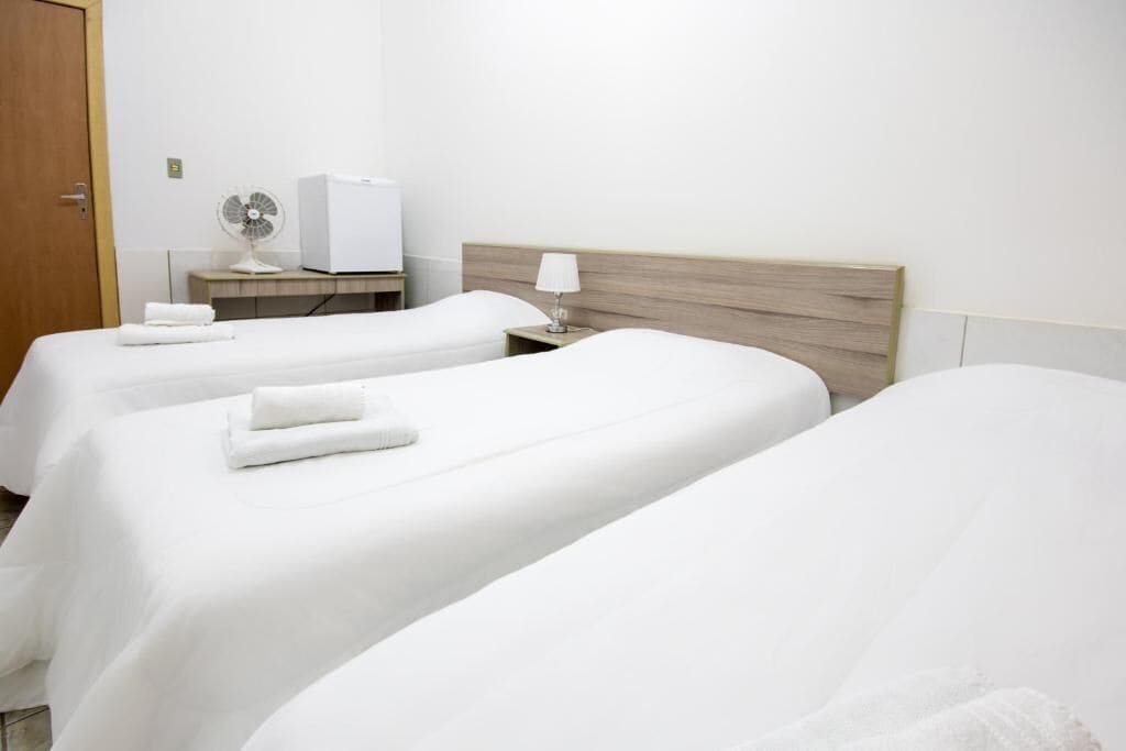 A hotel room with two single beds, each with a white bedspread and a folded towel. There is a wooden headboard, a bedside table with a lamp, and a white fan on the wall.