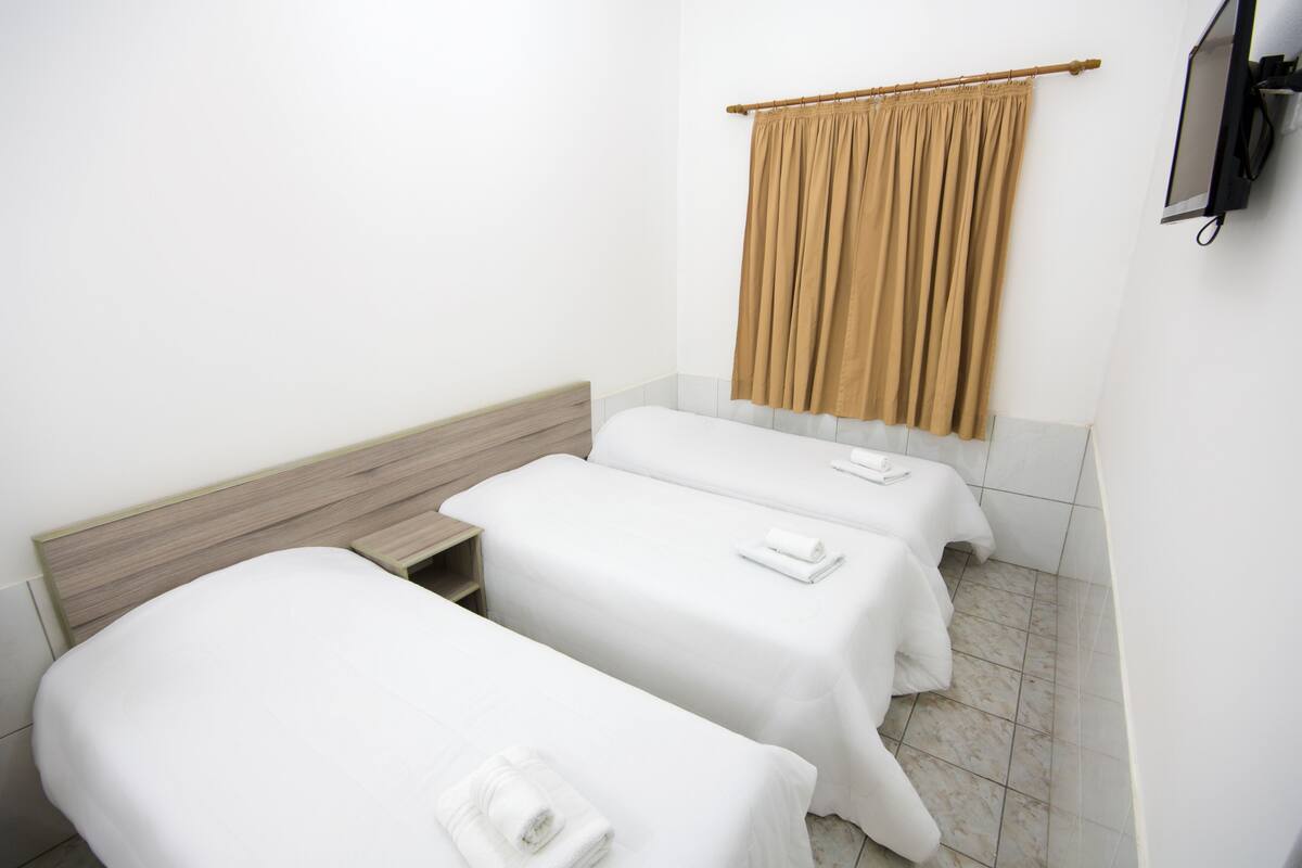 Ein Hotelzimmer mit zwei Einzelbetten, einem Holz-Kopfteil, einem beigen Vorhang und einem an der Wand montierten Flachbildfernseher.