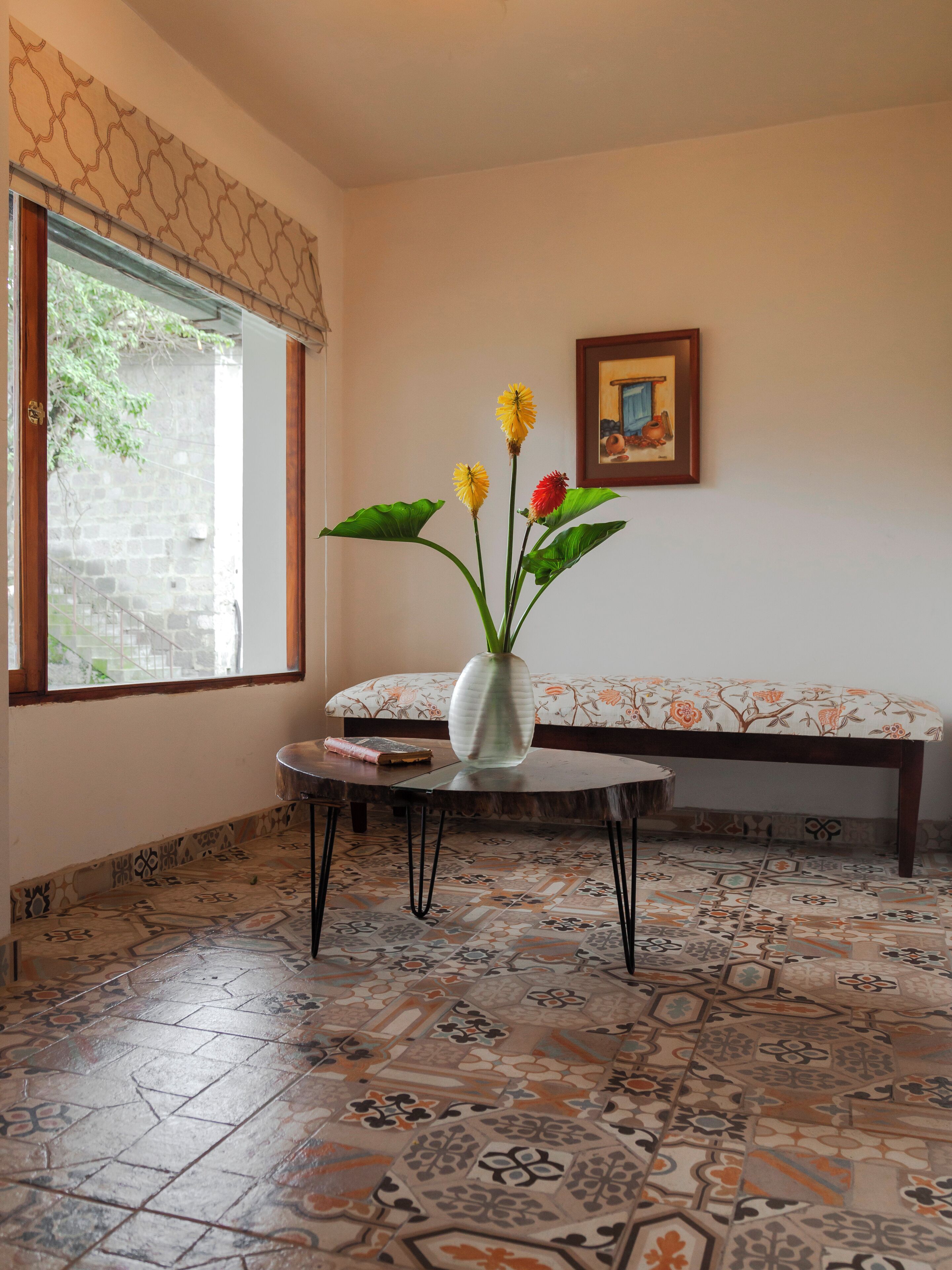 Una habitación con piso de baldosas estampadas, una mesa redonda con un florero, un banco y una ventana con vista a follaje.