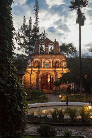 A stone building with a bell tower and arched windows, surrounded by lush greenery and a stone pathway. - Hacienda La Ciénega (Tanicuchi)