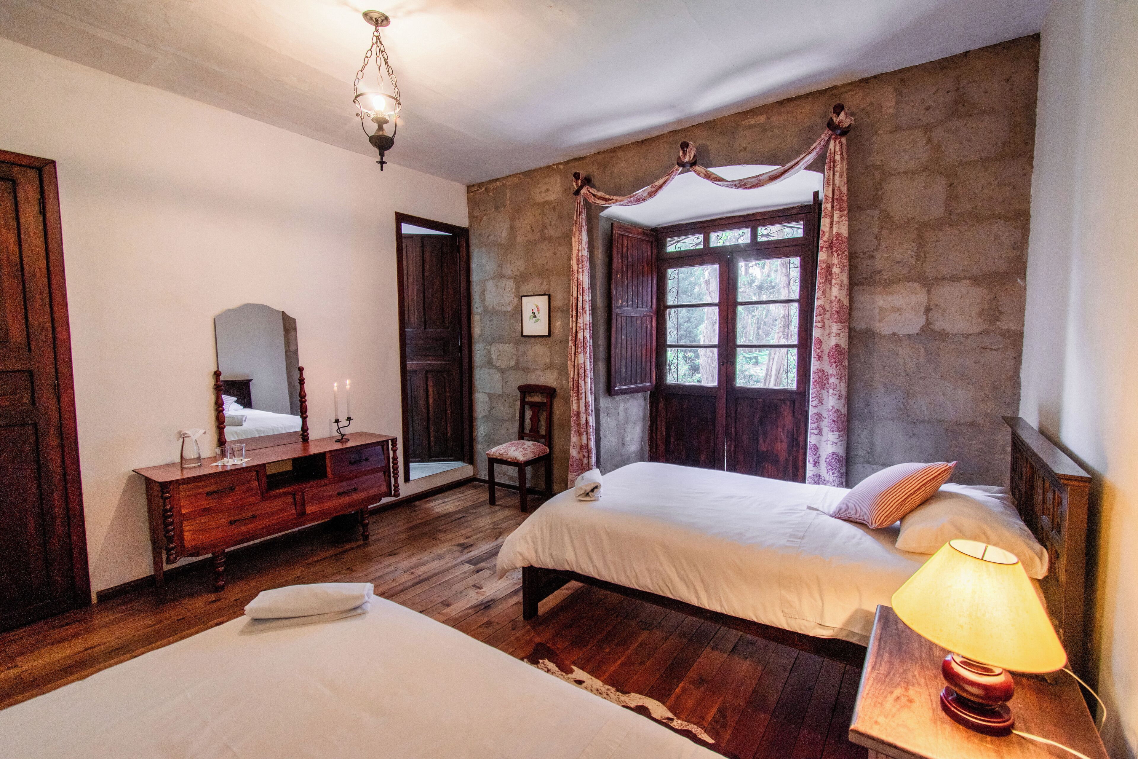 A bedroom with two beds, wooden floors, a dresser, a mirror, and a window with curtains.