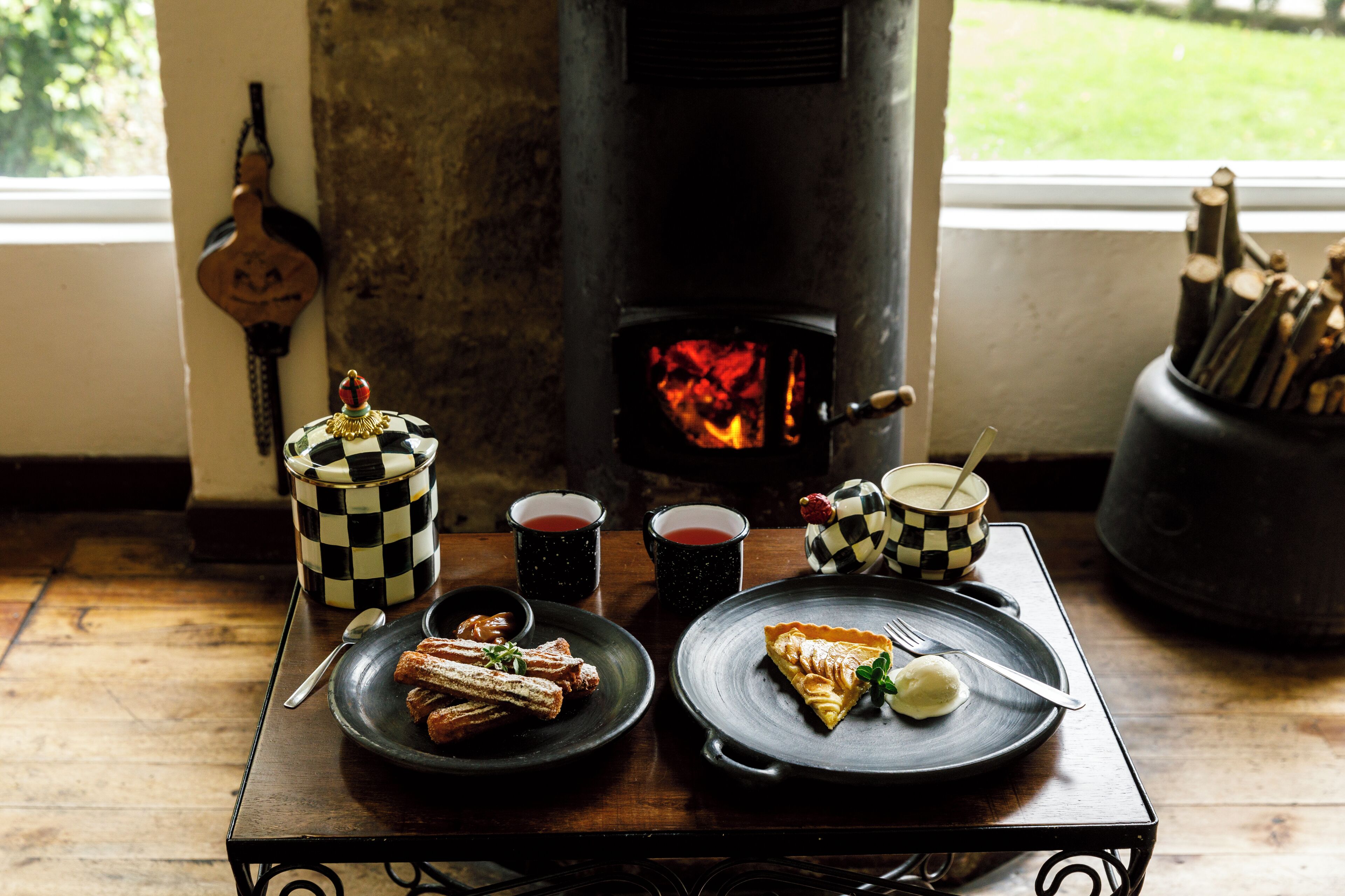 A rustic dining table with plates of pastries and a pie, two checkered mugs, and a wood-fired oven in the background.
