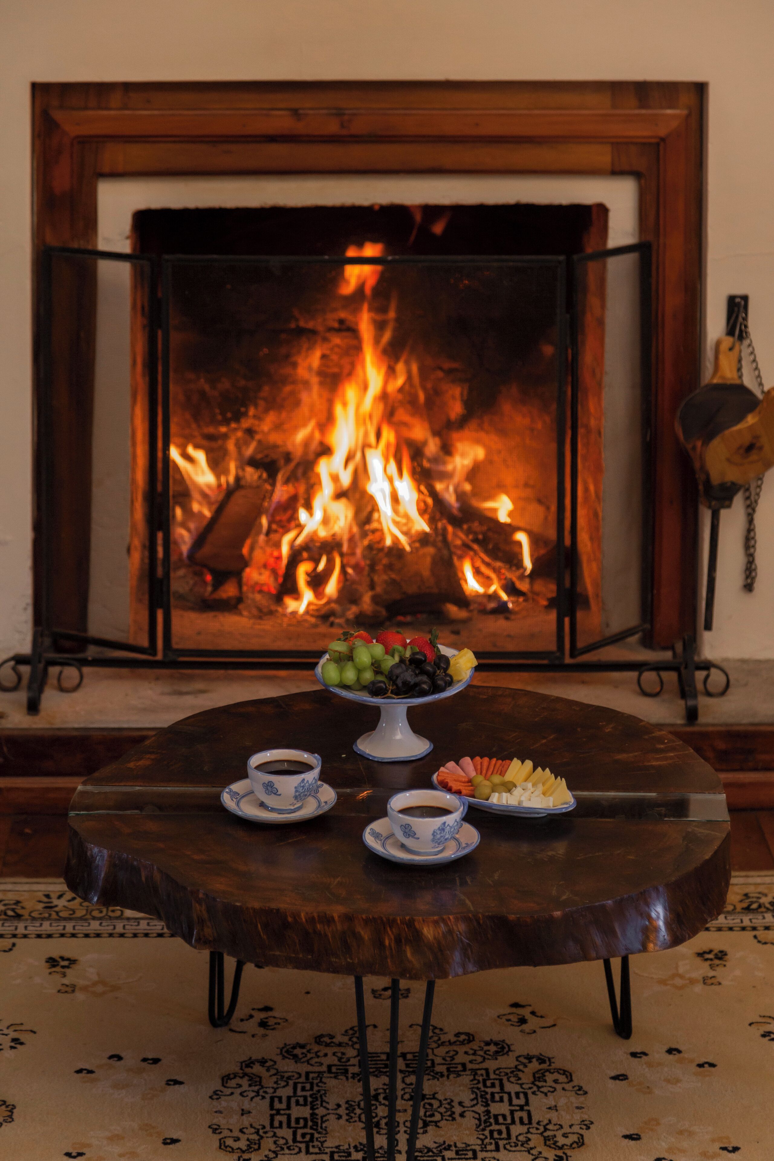 A cozy room with a fireplace, a round wooden table with a fruit bowl, cups, and a plate of snacks.