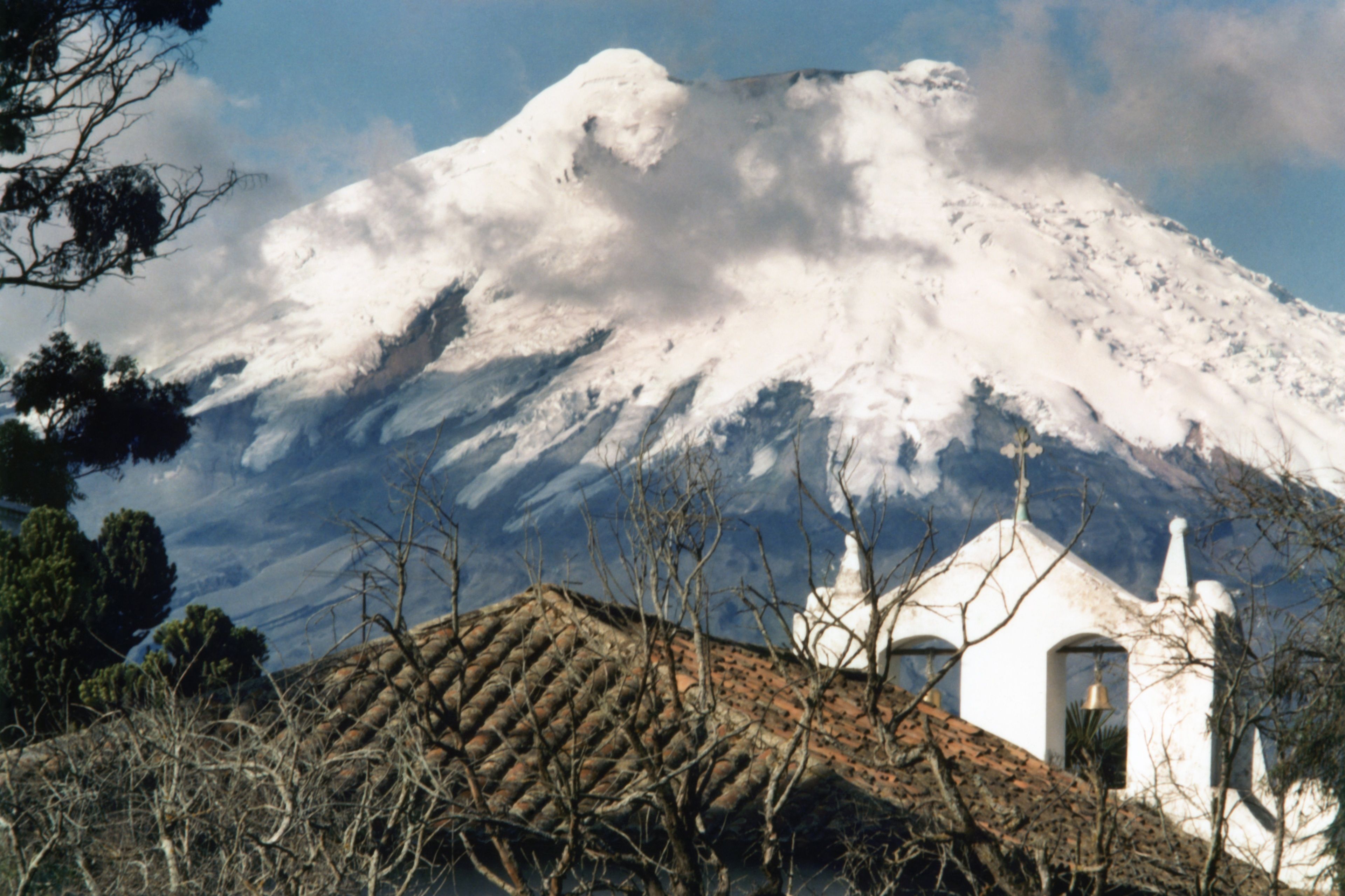 Una montaña con picos nevados, un edificio blanco con una torre de campanas y un tejado de tejas.